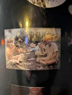 Street sticker The sticker depicts what appears to be three children outdoors. One child with bright orange hair is sitting upright. Another child is lying down with a dog nearby. A third child is sitting and holding something in their mouth, possibly a pipe or piece of grass. They are surrounded by grass and some cooking utensils, which suggests they may be camping or having a picnic.