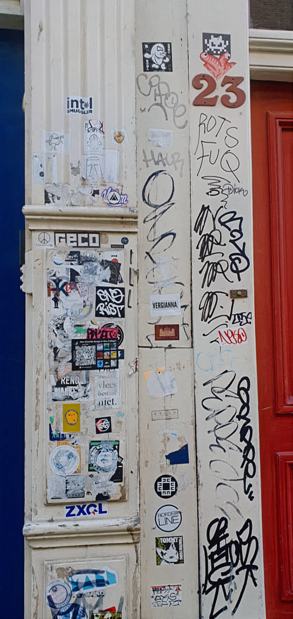 This image captures a corner of a building that's heavily decorated with various stickers and graffiti. The stickers range from simple designs to more elaborate artistic expressions, while the graffiti includes handwritten tags and abstract markings, creating a layered and textured appearance. The overall aesthetic suggests a vibrant urban environment where public spaces are used for self-expression and communication.