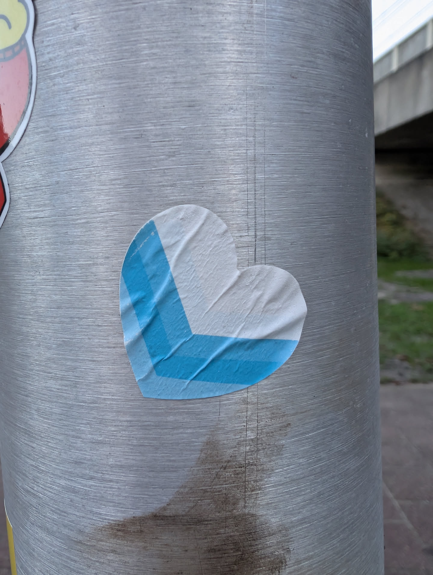 The sticker is heart-shaped and predominantly white, with a geometric design in bright blue. The blue forms an 'L' shape or a corner within the heart, with one segment running along the bottom curve and another extending upwards along the left side. The sticker appears to be made of paper and shows some creasing and wrinkling, indicating it has been applied to a textured surface. The edges are slightly irregular, and there are minor imperfections in the blue coloring.