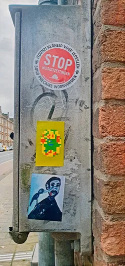 The sticker features an 8-bit style design in yellow, green, red, and blue, placed on a weathered metal post alongside other stickers. The post is situated next to a brick wall.