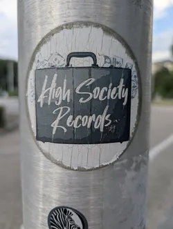 Street sticker Circular, weathered sticker featuring a black briefcase graphic with 'High Society Records' in white script. Faint illustrations of money bills appear in the white background. The sticker shows vertical cracking.