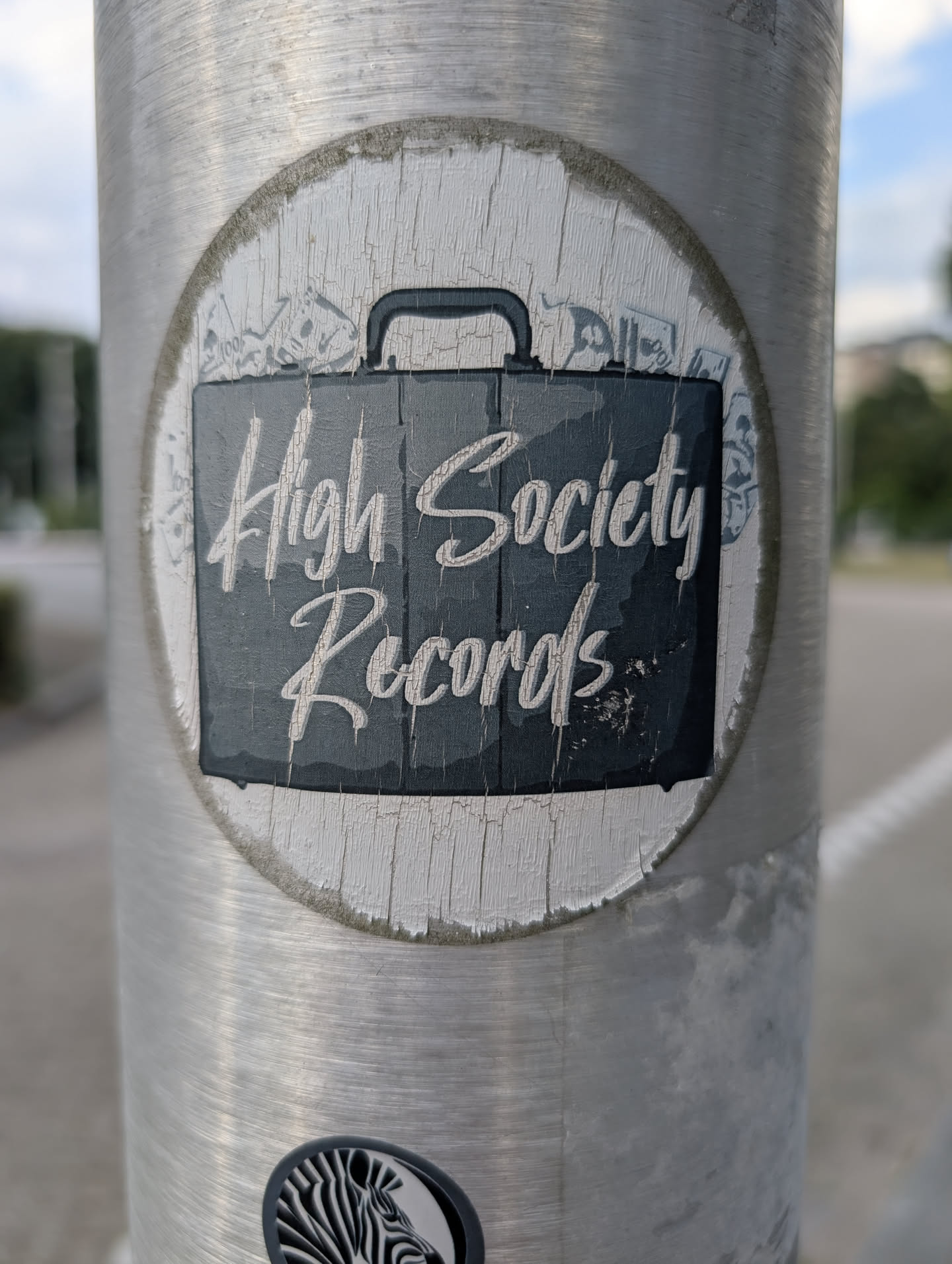 Circular, weathered sticker featuring a black briefcase graphic with 'High Society Records' in white script. Faint illustrations of money bills appear in the white background. The sticker shows vertical cracking.