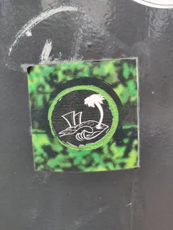A small sticker with a green and black design.  The main design element is a circle containing a stylized image of a palm tree on a small island with waves. The background of the sticker has a tie-dye or similar mottled effect in green and black.