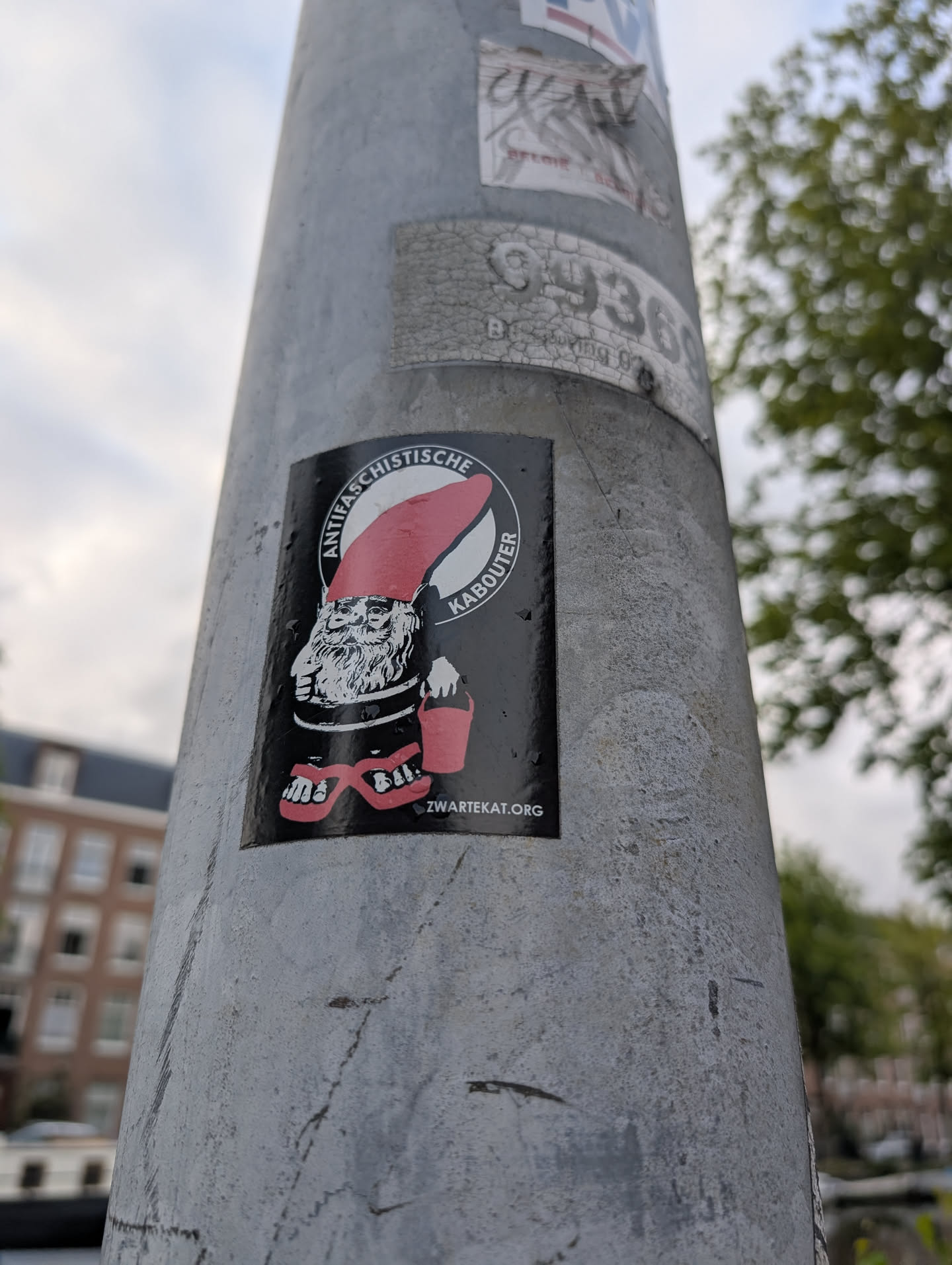 Street sticker The sticker is circular with a black background. In the center is a black and white illustration of a gnome wearing a large, pointed red hat. The gnome has a long white beard, bushy eyebrows, and appears to be wearing red flip-flops. It is holding a small red bucket in its left hand. Around the gnome, in a circular band of white text on the black background, are the words 'ANTIFASCHISTISCHE KABOUTER'. At the bottom center of the sticker, the text 'ZWARTEKAT.ORG' is visible.