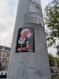 Street sticker The sticker is circular with a black background. In the center is a black and white illustration of a gnome wearing a large, pointed red hat. The gnome has a long white beard, bushy eyebrows, and appears to be wearing red flip-flops. It is holding a small red bucket in its left hand. Around the gnome, in a circular band of white text on the black background, are the words 'ANTIFASCHISTISCHE KABOUTER'. At the bottom center of the sticker, the text 'ZWARTEKAT.ORG' is visible.