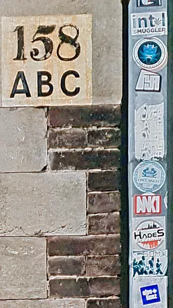 Street sticker This image showcases a variety of stickers affixed to a pole, beside a brick wall. The collection features diverse designs, each potentially representing different brands, messages, or artistic expressions. The pole and wall bear marks of weathering and age.