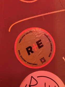Street sticker Round sticker with the text "R+E" in bold black letters, surrounded by the words "FREELANCE CREATIVE TEAM" repeated twice in a smaller font.  It has a QR code. The sticker appears to be beige or light brown with a red border.