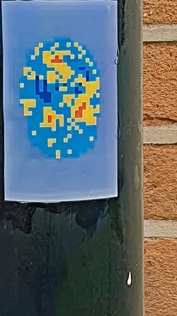 A pixelated sticker depicting a world map with blue and yellow continents, and a blue background. It is pasted on a black pole, next to a brick wall.