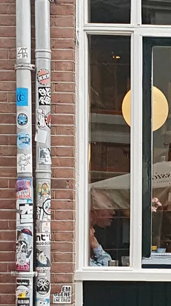 A view of a brick wall with a drainpipe covered in various stickers. Next to it, there's a window reflecting an interior scene with a person.