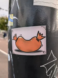 A sticker of a cartoon sausage with a crown on each end. The sausage is orange and the background is light pink. The sticker is placed on a dark gray pole.