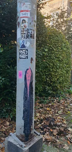 A tall, narrow sticker on a gray pole features a mostly black figure with pink highlights outlining the face and pink 'paint' dripping down. The figure appears somber or distressed. The pole also contains other stickers, leaves and bush around. 