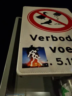 Street sticker A sticker featuring a stylized figure set against a backdrop of fire and smoke. The sticker is affixed to a traffic sign that prohibits feeding birds. The figure's design is abstract and bold, suggesting themes of protest or rebellion. 