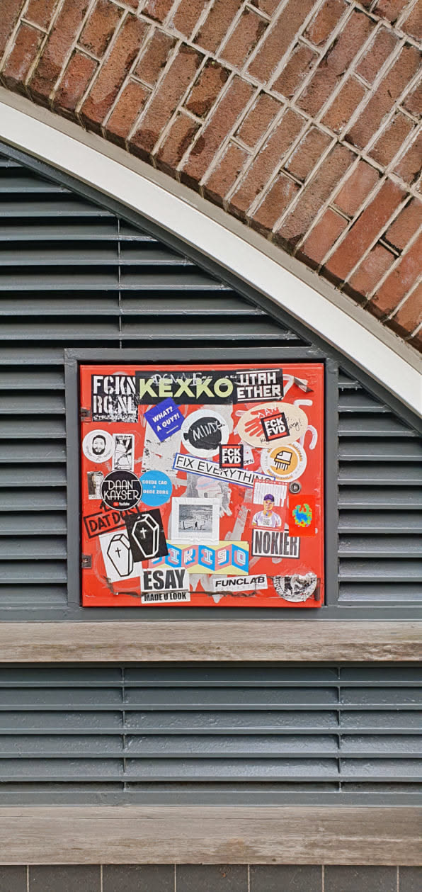 A red surface covered in various stickers, including designs with text and images. Some stickers appear to be from artists or brands. The stickers are densely packed, creating a collage-like effect. The surface is mounted on a wall with brickwork above and slats below. The stickers feature a range of styles, from hand-drawn to professionally printed.