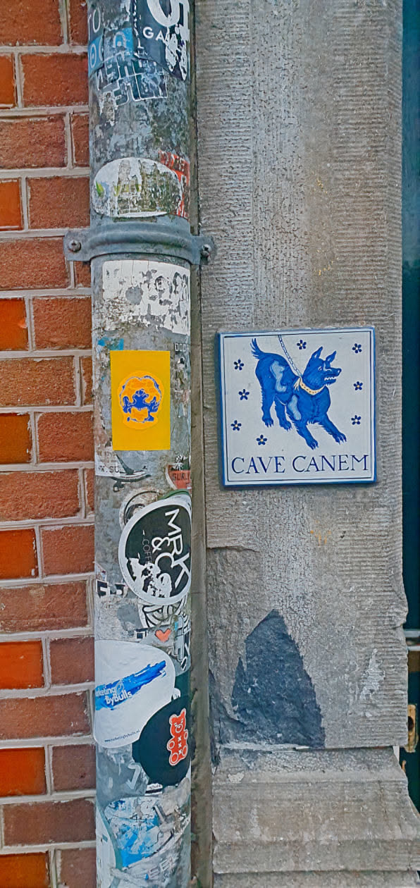The photo shows a yellow sticker affixed to a weathered metal pipe amidst other urban stickers and graffiti. The sticker displays an abstract design in blue and orange. To the right, mounted on a stone wall, is a traditional 'Cave Canem' sign depicting a dog. The scene suggests a blend of contemporary urban culture and classical motifs.