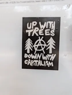Street sticker A black and white sticker with a hand-drawn style. It features the text "Up with Trees" over a stylized anarchy symbol and three evergreen trees, with the text "Down with Capitalism" below. 