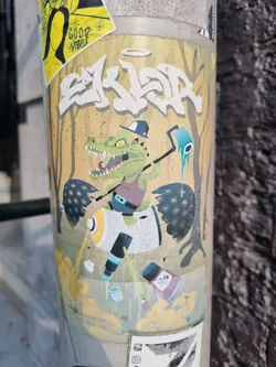 Street sticker This sticker features a cartoon crocodile wearing a hat and holding a paint roller, surrounded by graffiti elements and paint cans. The style is playful and colorful. The text at the top says "SKLDR" in graffiti style and there is a small halo above the word. At the bottom there is small text that is hard to read.  There are no social media links or QR codes visible in the image.