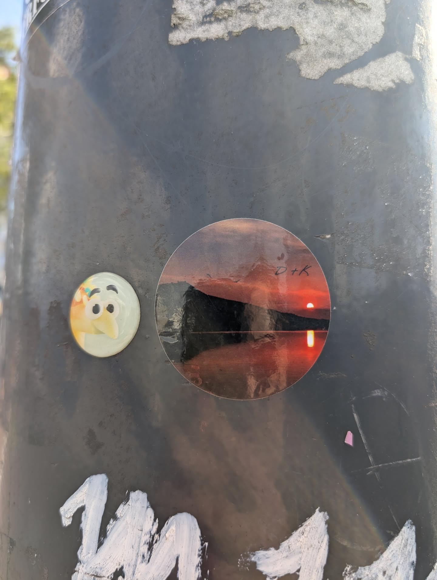 A round sticker affixed to a pole shows a vivid sunset over water, with a silhouetted mountain range in the background. The sun reflects intensely on the water's surface, creating a dramatic visual. A very small text "D+K" is written on the top right of the sticker. The sticker is circular.