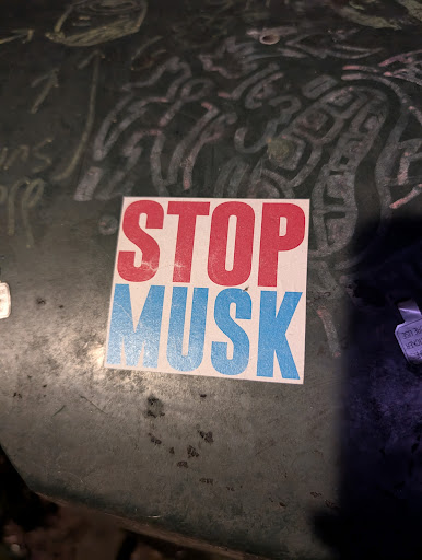 A square sticker with the words "STOP MUSK" in bold capital letters. "STOP" is in red, while "MUSK" is in blue. The sticker is placed on a dark surface with some smudges and inscriptions around it. The overall impression is a political statement against Elon Musk.