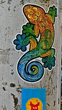 Street sticker This sticker depicts a lizard with an artistic design. The head is yellow and transitions to orange, then green, and finally blue at the tail. The pattern is a swirling design that is both organic and geometric. The text 'DHMO' is visible near the tail of the lizard.