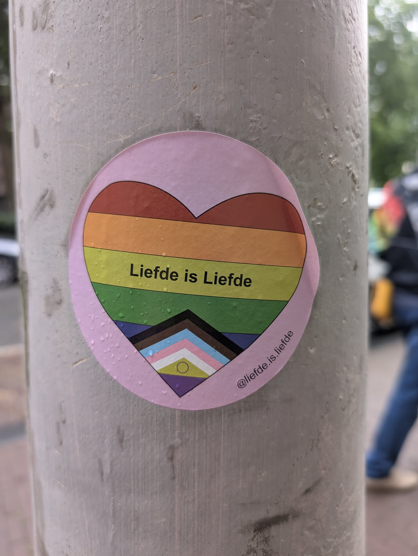 A circular pink sticker with a light pink border, featuring a heart design at its center. The heart is divided into horizontal stripes representing the rainbow flag, with additional stripes from the Progress Pride flag at the bottom. The text "Liefde is Liefde" is prominently displayed in black within the heart. There are small water droplets visible on the sticker's surface. The sticker is adhered to a textured, light gray cylindrical surface, likely a pole.