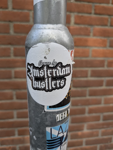 The sticker has a white background with black text outlined in white. The text 'Crown by' is at the top in a stylized font, followed by 'Amsterdam Busters' in a larger, gothic-style font. The sticker is affixed to a metal pole with a brick wall visible in the background.