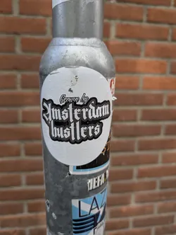 Street sticker The sticker has a white background with black text outlined in white. The text 'Crown by' is at the top in a stylized font, followed by 'Amsterdam Busters' in a larger, gothic-style font. The sticker is affixed to a metal pole with a brick wall visible in the background.