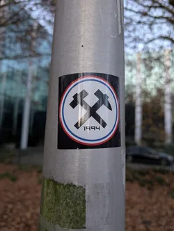 The sticker is circular with a black background. It features a white circle in the center, bordered by a thin red line and then a thin blue line. Inside the white circle, two black, stylized hammers are crossed. The hammer on the left has its head pointing upwards and to the left, while the hammer on the right has its head pointing upwards and to the right. Below the crossed hammers, the number '1994' is printed in black, blocky digits.