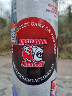 Street sticker A round sticker with a red and black design featuring a bear logo and the text 'Amsterdam Lacrosse'. The text 'Fastest Game on Two Feet' is also present on the sticker.  A QR code is partially visible to the left of the main design. The sticker is affixed to a pole.