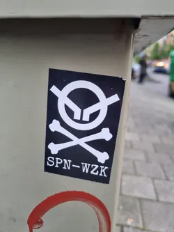 A black and white sticker with a stylized skull and crossbones graphic. The text 'SPN-WZK' is printed below the graphic. The sticker is affixed to a light beige surface outdoors.