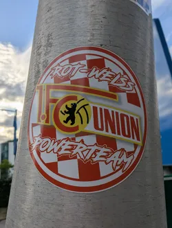 Street sticker A circular sticker featuring the 1. FC Union Berlin logo over a red and white checkered background. The words 'ROT-WEISS' and 'POWERTEAM' are written in a stylized white font at the top and bottom. The central club logo displays a black bear in a yellow circle alongside the text '1. FC UNION'.