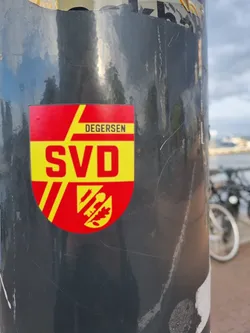 Street sticker A red and yellow sticker featuring the logo of a German football club, Degersen SVD. The logo is a shield with the club's initials and a small emblem. The sticker is affixed to a dark grey pole.
