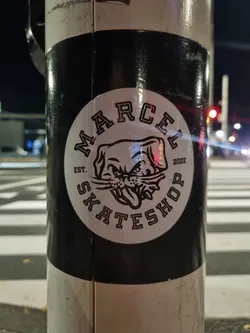 Street sticker A black and white circular sticker featuring a dog mascot with the text 'Marcel Skateshop' and 'Est. 2022'. The sticker is affixed to a pole outdoors at night.