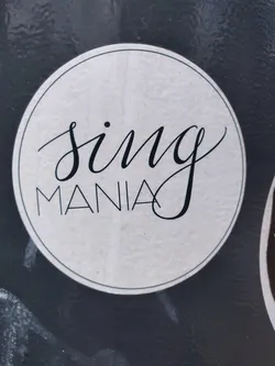 Street sticker A round sticker with the text "Sing Mania" written in an elegant, stylized font. The text is in black on a white, textured background, which is circled by a thin black border.