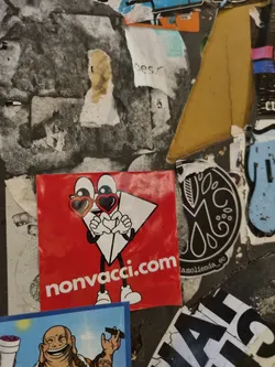 A red square sticker features a cartoon character resembling a paper airplane with heart-shaped sunglasses. The character is holding its hands in a heart shape. The website nonvacci.com is printed at the bottom. The sticker is affixed to a wall amongst other stickers.