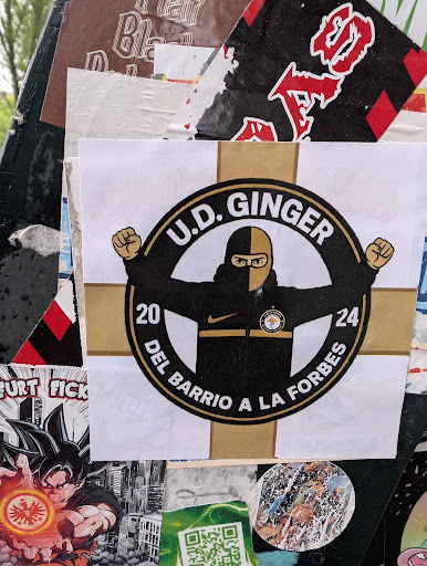 The central sticker showcases a white background adorned with a black and gold crest design. It features the text "U.D. GINGER" at the top, with an image of a figure wearing a black mask and jersey, fists raised. The figure is encircled by the text "20" and "24" at either side. The text "DEL BARRIO A LA FORBES" is at the bottom. The overall aesthetic suggests a soccer fan or supporter group.