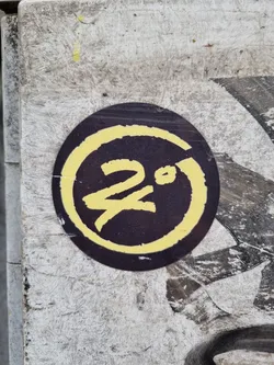 Street sticker A round, dark-colored sticker with a yellow circle and stylized characters in the center. The sticker appears to be affixed to a weathered surface. The central characters are not easily identifiable.
