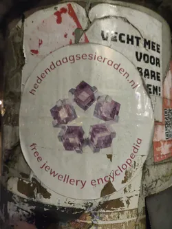 Street sticker A round sticker with a pink design of what looks like crystals or gemstones arranged in a star shape. The text on the sticker says "Free Jewellery Encyclopedia" and also shows a website address: hedendaagssieraden.nl
