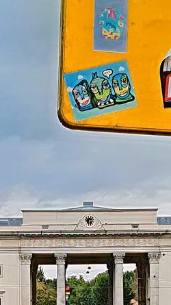 Street sticker A sticker with three stylized figures on a blue background, adhered to a yellow surface. The figures have unique facial features and one has a speech bubble that says 'HELLO!!'.