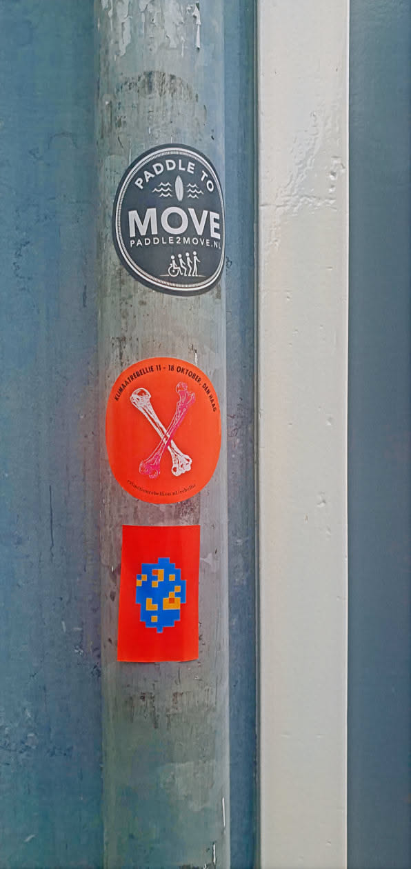 A photo of three stickers on a metal pole. The top sticker is round and black with the words "PADDLE TO MOVE PADDLE2MOVE.NL" and a graphic of a surfer and some figures in wheelchairs. The middle sticker is round and orange with the words "KLIMAATREBELLIE 11 - 18 OKTOBER, DEN HAAG" and an image of crossed bones. The bottom sticker is square and red with blue and yellow pixel art.