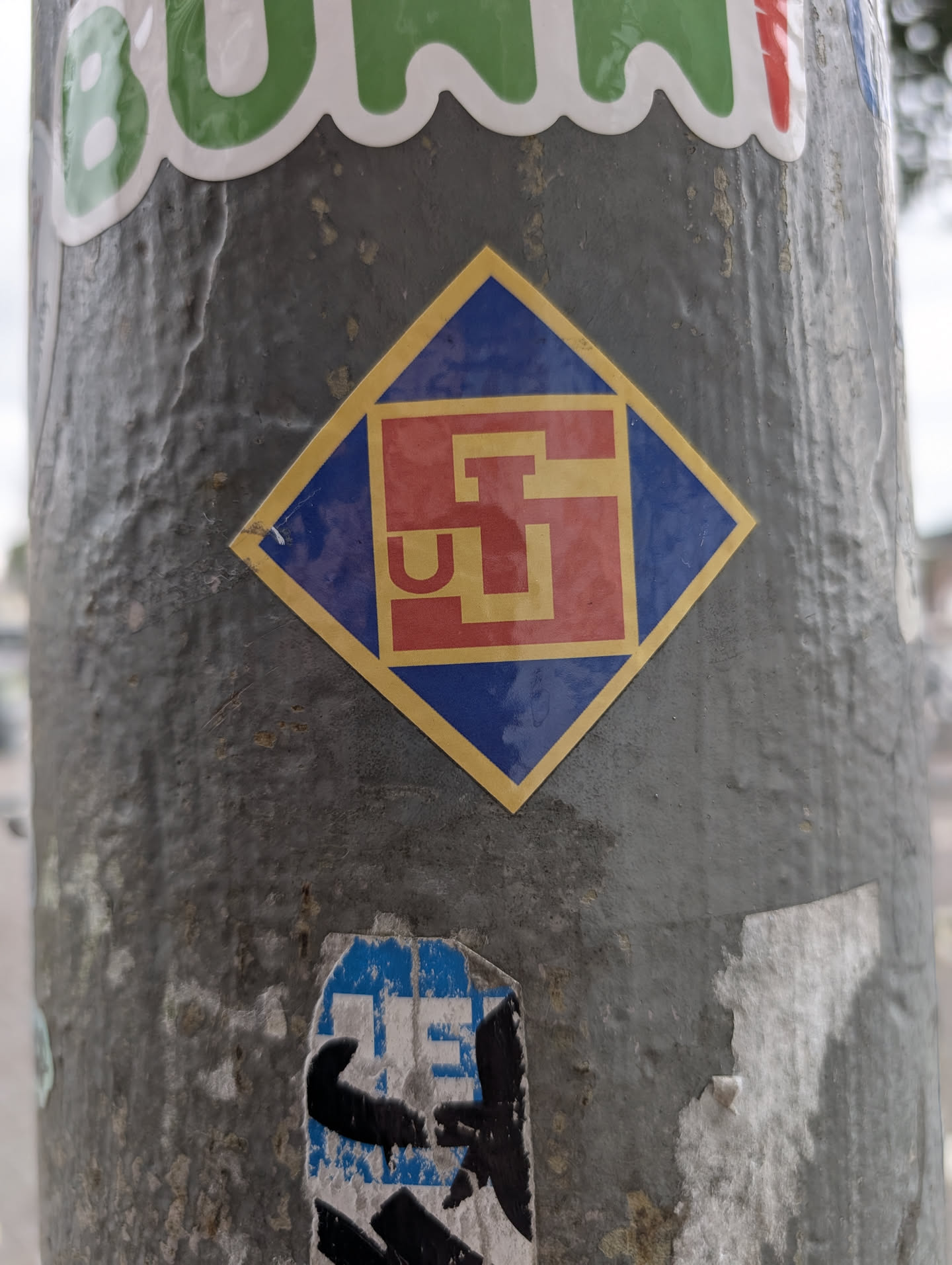 The sticker is square-shaped but oriented like a diamond. It features a thick yellow border. Inside this border, there is a solid blue square. Within the blue square, a red square is placed slightly off-center. The central element is a stylized, abstract symbol rendered in a bright yellow, resembling a simplified 'G' or a stylized 'H' with a curved base, enclosed within the red square. The overall design is clean, modern, and bold, with high contrast between the colors.