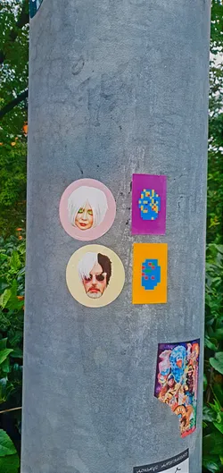 A variety of stickers adorn a pole. The top left sticker is a round portrait of a person with white hair on a pink background. Below that is another round portrait of a person with sunglasses and black and white hair. To the right of these are two rectangular stickers with 8-bit style artwork, one on a purple background and the other on orange. At the bottom is a sticker with a collage of characters and designs, and a small bit of text.