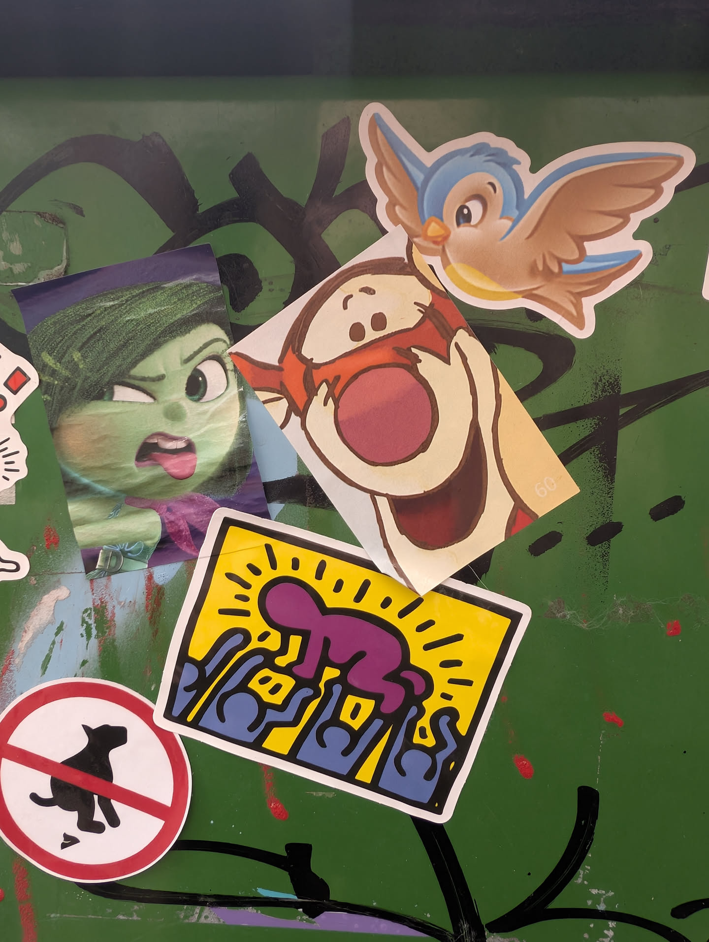 The sticker comprises multiple individual stickers layered on top of each other. Prominently featured is a Keith Haring-style graphic with a purple figure over blue figures against a yellow background. To its left is a circular sticker with a black dog silhouette in a 'no pets' sign style. Above these, a sticker depicts the character Disgust from Pixar's 'Inside Out' with a green face and a disgusted expression. To the right of Disgust is a sticker of Tigger from Winnie the Pooh, with his mouth wide open and smiling. A small blue bird is also visible, positioned above Tigger.