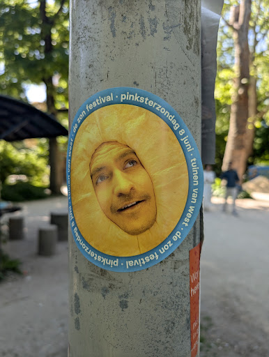 A round sticker on a pole shows a man's face framed by a bright yellow sun shape. The sticker advertises a festival, with text circling the edge. The background is a blurry outdoor scene with trees and a path.