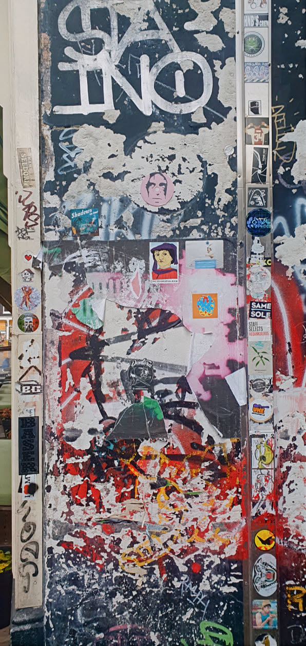 The wall is covered in a variety of stickers and graffiti, creating a layered and textured effect. Some stickers are easily identifiable, while others are obscured by the overlapping design. The overall impression is one of urban decay and artistic expression.