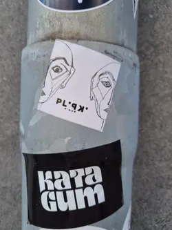 A small white square sticker with a black and white line drawing of two faces looking in opposite directions. The text 'PL·9K·' is visible below the drawing.  The sticker is attached to a metal pole, next to another sticker reading 'Kara Gum'.