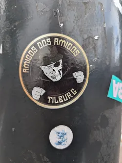 Street sticker A round sticker featuring a skull-faced man in a hat, with the text "Amigos Dos Amigos Tilburg" around him. The sticker has a dark background with gold lettering. 