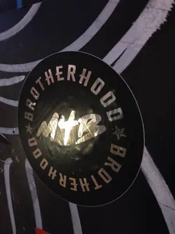 Street sticker A black round sticker with the text "Brotherhood" written in a circular pattern around a central logo that appears to say "MTB". The text and logo are in a metallic gold color.