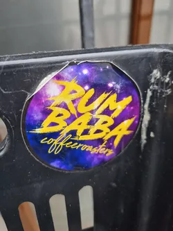Street sticker A round sticker with a purple and blue cosmic background. The text \"RUM BABA\" is written in yellow with a graffiti style. Underneath, it says \"coffeeroasters\" in a smaller font. The sticker is attached to a dark-colored surface.