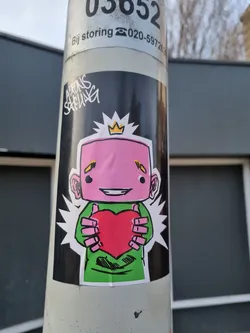 A sticker featuring a pink cartoon character with a crown holding a red heart. The character is wearing a green shirt. The sticker is placed on a pole.