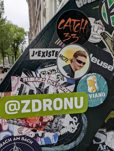 A photograph of a street sign densely covered with various stickers. Notable stickers include one with the text "CATCH 33", another with "J'EXISTE", and a green sticker displaying "@ZDRONU".
